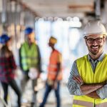 Photo of business people group on meeting and presentation in bright modern office with construction engineer architect and worker looking building model. Front view of smiling architect in helmet against his three colleagues.